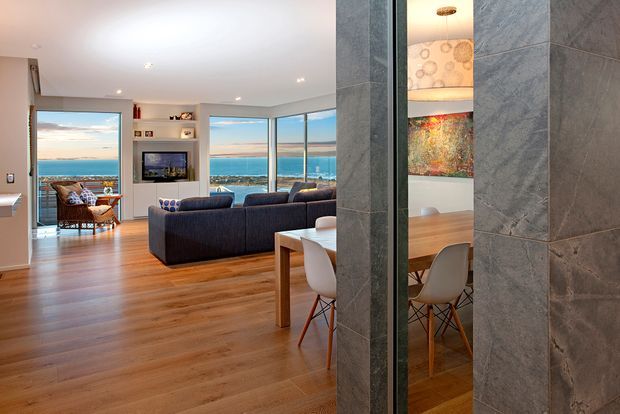 Open-plan contemporary living and dining area with warm timber flooring, a large dark sectional sofa, recessed ceiling lighting, and floor-to-ceiling glazing overlooking a coastal waterfront. A dining table and chairs are visible through a central opening, with a tiled feature wall and built-in television joinery.