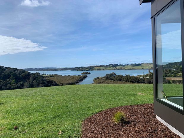 Exterior view from a landscaped grassy site toward a calm coastal inlet, with wooded shoreline and rolling rural land beyond. The right side shows the corner of a contemporary building with dark cladding, large floor-to-ceiling glazing, and a projecting roof edge. A mulched planting bed borders the building.