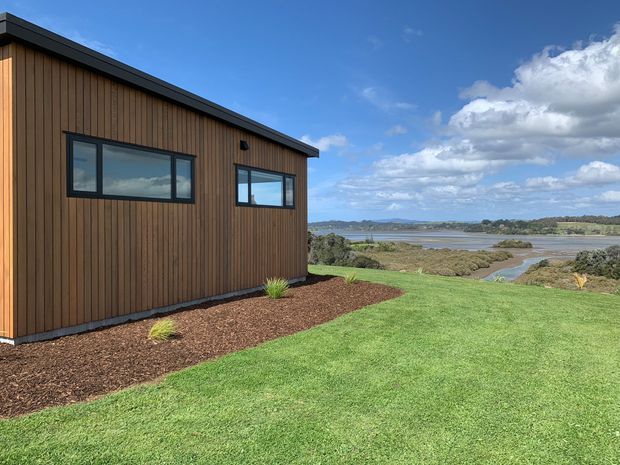 Exterior view of a compact modern building with dark timber vertical cladding, black-framed rectangular windows, and a low sloping roof. The building sits beside a landscaped lawn and mulched garden beds, with a broad tidal or coastal water landscape visible beyond.