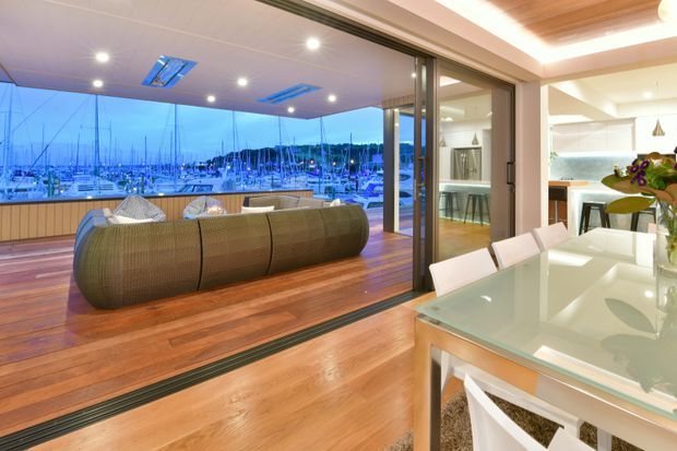 Open-plan contemporary interior overlooking a marina, with a polished timber floor, large sliding glazed doors, a curved upholstered sofa in the living area, and a bright white kitchen and dining space. Recessed ceiling lighting and integrated cabinetry create a refined, modern fit-out.