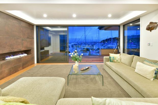 Contemporary living room with a large beige sectional sofa and ottoman, glass coffee table, and a linear built-in gas fireplace set into a brown stone feature wall. Full-height sliding glass doors open to a waterfront marina view and outdoor deck. A recessed ceiling with integrated downlights creates a polished, relaxed atmosphere.