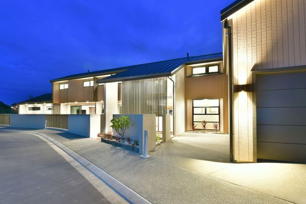 A dusk exterior view of a contemporary multi-unit residential development, showing clean-lined two-storey homes with illuminated windows, vertically ribbed facade panels, dark metal pitched roofs, garages, covered entries, and paved driveways. The architecture is presented in a quiet suburban streetscape with strong warm lighting against a deep blue sky.