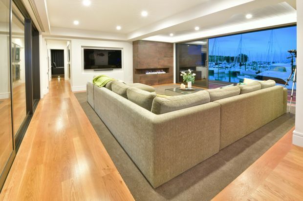 A spacious contemporary living room with polished timber flooring, a large neutral sectional sofa, recessed ceiling lights, built-in television cabinetry, and a brick feature wall with fireplace. Full-height glazing opens views toward a marina, while dark-framed interior glazing and warm wood finishes create a refined, inviting atmosphere.