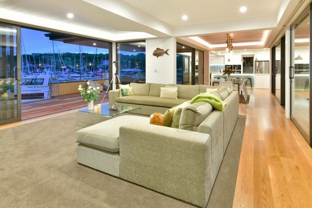 A spacious, open-plan living room with a large L-shaped upholstered sectional sofa, neutral carpet, and warm timber flooring. Full-height glazing opens to a marina-facing deck, while recessed ceiling lighting and a timber-accented ceiling detail create a polished contemporary interior. The kitchen and dining area are visible in the background.