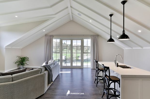 Bright open-plan attic-style living and kitchen space with a vaulted white ceiling, exposed painted beams, dark timber flooring, a light grey sectional sofa, and a white island with bar stools. Large glazed doors open to a balcony or exterior view, while black pendant lights provide contrast.