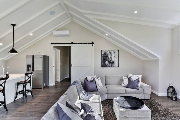 Bright open-plan loft-style living area with a vaulted white-lined ceiling and exposed beams, light grey sectional seating, dark timber-look flooring, a white kitchen peninsula with bar stools, stainless-steel refrigerator, and a black sliding barn door. Neutral furnishings with purple accent cushions create a contemporary, inviting interior.