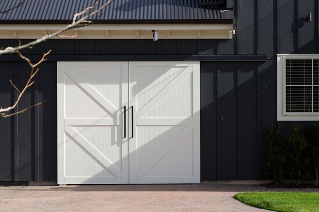 Close-up exterior view of a dark-clad building facade featuring large white barn-style double doors, a corrugated metal pitched roof, and a small white-framed window with security bars. A paved driveway and narrow lawn edge are visible in the foreground.