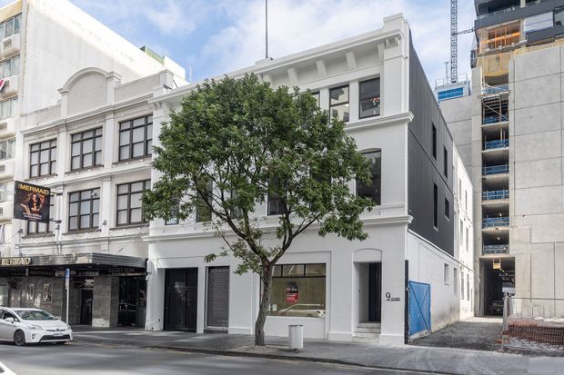 Street-level exterior view of a narrow, multi-storey white rendered commercial or mixed-use building in an urban setting. The facade features tall dark-framed windows, classical-style mouldings, decorative cornices and pilasters, with neighbouring heritage-style and contemporary apartment construction visible on either side.