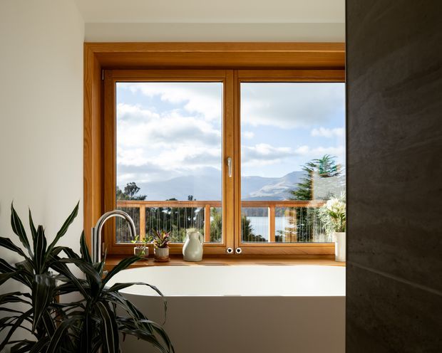 Bright interior view focused on a timber-framed double window above a deep white sill or counter. A tall leafy indoor plant and small potted plants sit near the window, while a curved chrome faucet and partial basin suggest a kitchen or utility area. Through the glazing, a balcony railing, water, and mountainous landscape are visible. The space has clean white surfaces, warm timber joinery, and a dark textured wall or partition at right.