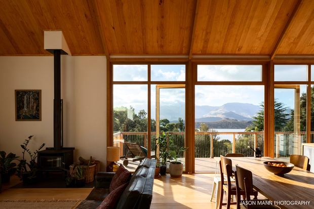 Warm, timber-lined open-plan living and dining space with a wood-burning stove, leather sofa, timber dining table, and extensive floor-to-ceiling glazing opening to a deck. The windows frame a scenic mountain and water view, while indoor plants and natural materials create a relaxed, light-filled atmosphere.