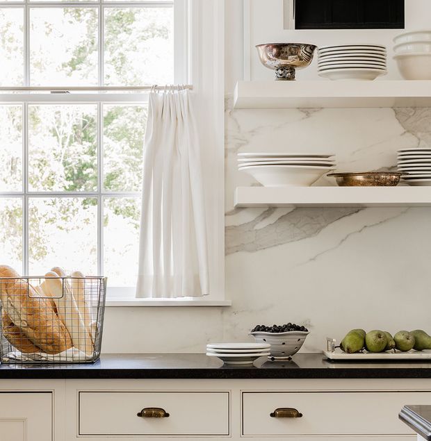 Bright, refined kitchen vignette with a large multi-pane window and white curtain beside open marble-look shelving displaying white and metallic ceramics. A dark countertop runs above cream cabinetry with brass cup pulls, while fruit and bread baskets add understated warmth.