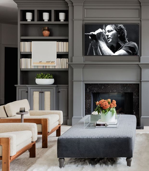A refined living room with dark grey built-in shelving and panelled fireplace cabinetry, a wall-mounted television, upholstered timber-framed armchairs, and a large grey ottoman coffee table. The room has a sophisticated monochromatic palette with warm wood accents and a floral arrangement.