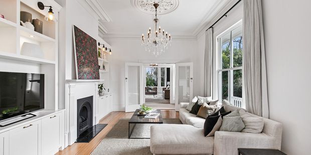 Bright, elegant living room with white built-in cabinetry, a television, traditional fireplace surround, ornate ceiling rose and cornices, chandelier, tall sash-style windows with full-length curtains, timber flooring, and a large neutral sectional sofa. The room opens through glazed double doors to another interior space and is styled in a refined traditional manner.