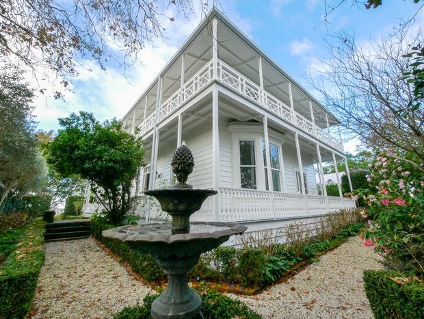 Exterior view of a grand white two-storey house with extensive decorative verandahs, ornate balustrades, slender columns, and a steep gabled roof. The landscaped garden includes gravel paths, clipped shrubs, flowering plants, and a dark ornamental fountain in the foreground.
