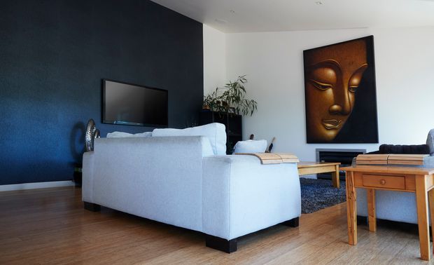 A modern living room viewed from behind a light upholstered sofa, with warm timber flooring, a dark navy feature wall, wall-mounted television, timber coffee and side tables, and a large Buddha artwork. The room has a clean, contemporary arrangement with strong contrast between the dark feature wall and light furnishings.
