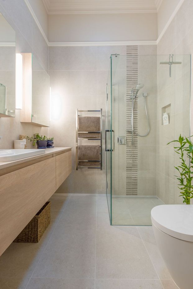 Bright contemporary bathroom with a walk-in glass shower, wall-mounted timber vanity, large pale floor and wall tiles, chrome fixtures, a heated towel rail, and a white toilet. Subtle greenery and woven storage add natural accents.