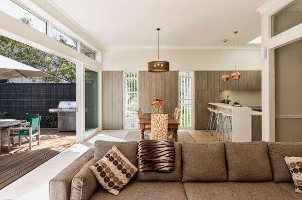 Bright open-plan living area with a large brown sectional sofa in the foreground, a dining table and chairs beyond, and a compact white kitchen with an island and stools. Full-height glazing opens onto a timber outdoor deck with a barbecue. The room features light wood cabinetry, white walls and ceilings, decorative pendant lighting, and strong indoor-outdoor connection.