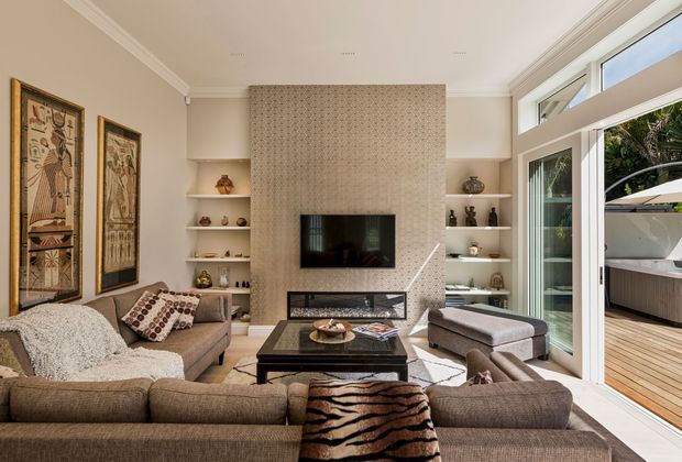 A refined living room with a large sectional sofa arranged around a dark coffee table and a textured stone or tile fireplace wall with a built-in television and linear fireplace. Built-in open shelving displays decorative objects, while framed artwork hangs on the side wall. Full-height glazed sliding doors open to a timber deck and outdoor area, bringing in abundant natural light.