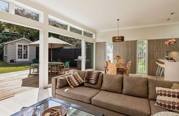 A bright open-plan living room with a large brown sectional sofa, patterned cushions, glass coffee table, and dining area beyond. The room opens through extensive glazing and sliding doors onto a timber deck and landscaped yard with a small garden studio. Vertical timber wall panelling, white-painted walls and trim, high clerestory windows, pendant lighting, and a compact white kitchen/bar area create a warm contemporary interior.