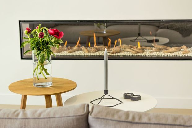 A contemporary living room vignette featuring a long horizontal built-in linear fireplace set into a white wall, with decorative stones and multiple small flames. A large mirror above the fireplace reflects the room. In the foreground are a light upholstered sofa, round timber side table with a glass vase of red flowers, a white round coffee table, and a slim modern candleholder.