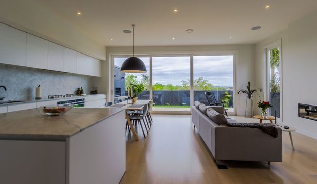 Open-plan contemporary kitchen and living area with white flat-panel cabinetry, a large stone island, timber flooring, dining table, and grey sofa. Full-height glazed doors open to a balcony or terrace with distant greenery and water or sky views. A minimalist linear fireplace is built into the right wall.