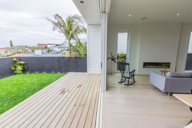Bright contemporary indoor-outdoor living area opening onto a timber deck and small lawn. The interior has pale timber flooring, white walls and ceiling, recessed lighting, a linear fireplace, a rocking chair and a light upholstered sofa. Large open glazing connects the living room to the landscaped outdoor space, with suburban rooftops visible beyond the fence.