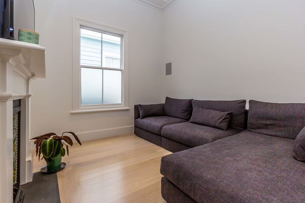 A bright, compact living room with a large dark charcoal sectional sofa, pale timber flooring, white walls and trim, a traditional white fireplace, and a single window. A potted plant adds a small natural accent.