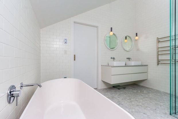 Bright contemporary bathroom with a freestanding white bathtub in the foreground, white subway-tiled walls, a floating double vanity with two basins, round mirrors, pendant lights, a heated towel rail, and a glass shower enclosure. The room has a clean, refined palette of white, pale grey, and muted green accents.