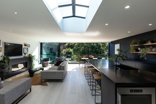 Bright contemporary open-plan living, dining and kitchen space with a large central island, dark cabinetry, integrated appliances, grey upholstered seating, a fireplace and expansive glazing to greenery. A dramatic linear skylight brings strong daylight into the room, while recessed lighting and open shelving add a refined modern finish.