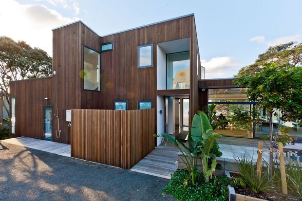 Exterior view of a contemporary multi-level home clad predominantly in vertical dark timber, with asymmetrical rooflines, large glazed openings, a timber privacy fence, covered entry, and landscaped planting around a deck and paved outdoor area.