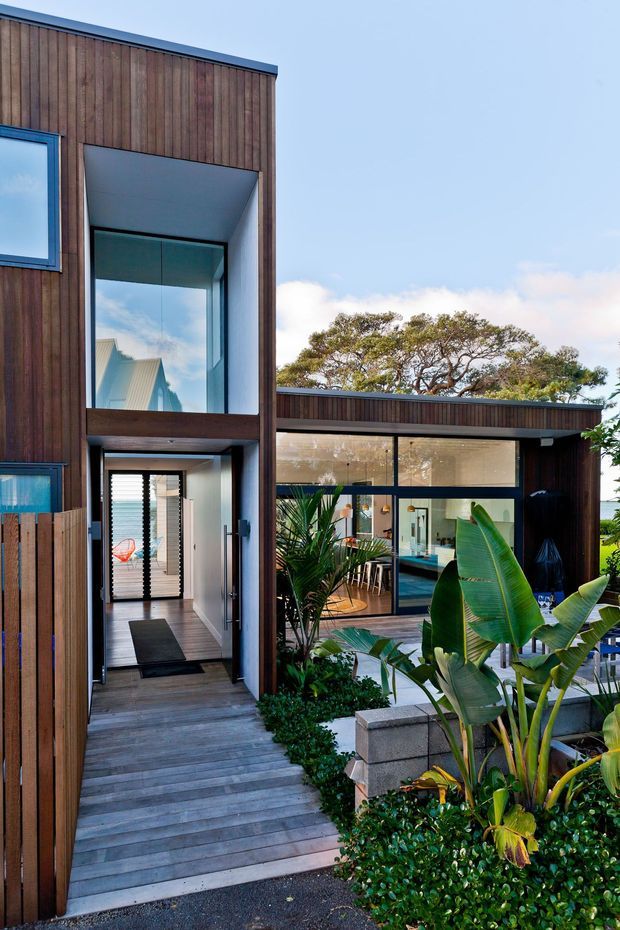 Exterior view of a contemporary multi-level home with dark timber cladding, expansive glazing, a recessed entry, timber walkway, and lush tropical landscaping. The open-plan interior and dining area are visible through the windows, with a low flat-roofed wing extending to the right.
