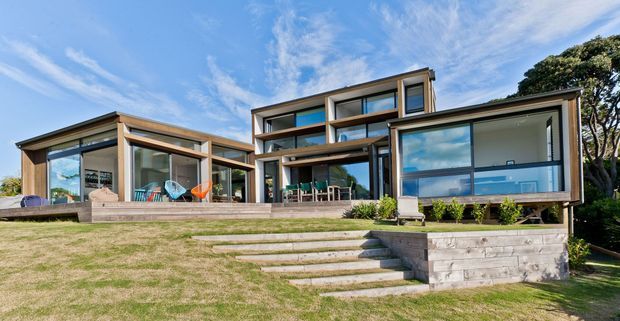 Modern multi-level residential home set into a gently sloping landscaped site, featuring extensive floor-to-ceiling glazing, timber and dark metal framing, projecting volumes, broad terraces, and a stepped timber retaining wall. The composition highlights the facade, outdoor deck, and connection between the interior and lawn.