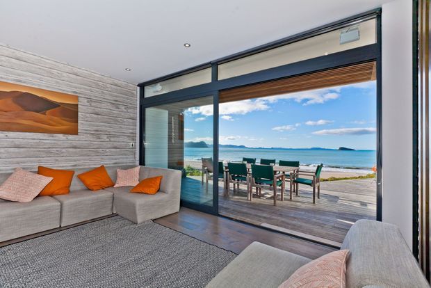 A bright contemporary living room opens directly onto a waterfront deck through large black-framed sliding glass doors. The room features grey sectional seating, orange and blush cushions, a textured grey rug, a weathered timber feature wall, and ocean views with an outdoor dining setting beyond.