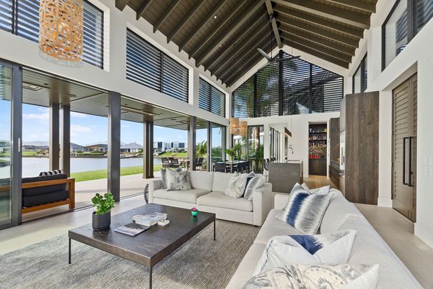 A spacious, light-filled contemporary living room with a large sectional sofa arrangement, dark timber coffee table, and expansive floor-to-ceiling glazing overlooking a landscaped waterfront setting. The room features a dramatic exposed pitched timber ceiling with exposed rafters, ceiling fans, dark louvred sun-control screens, timber wall panelling, and a compact bar or kitchenette area at the rear.