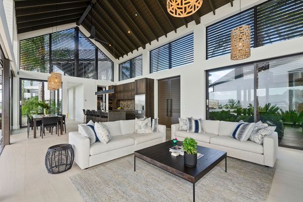 Bright open-plan living room with a large white sectional sofa, dark rectangular coffee table, textured area rug, and woven pendant lights. The space has high ceilings with exposed dark timber rafters, extensive glazing with horizontal louvre blinds, white walls, and a kitchen and dining area visible in the background.