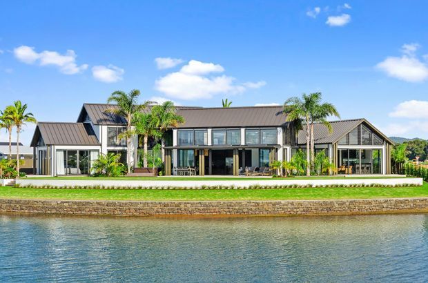 Wide exterior view of a large contemporary waterfront residence with a symmetrical, low-profile composition, dark standing-seam metal gabled roofs, extensive floor-to-ceiling glazing, and multiple connected wings. The landscaped lawn and palm planting sit directly beside a stone-edged canal or waterway under a bright blue sky.