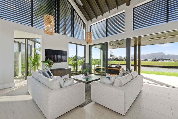 Bright, spacious contemporary living room with soaring exposed timber-beam ceilings, extensive floor-to-ceiling glazing, black horizontal blinds, pale tiled flooring, and two light upholstered sofas arranged around a coffee table. A television and low built-in console sit against the wall, while the glazing opens to a sunny landscaped setting with distant mountains and water beyond.