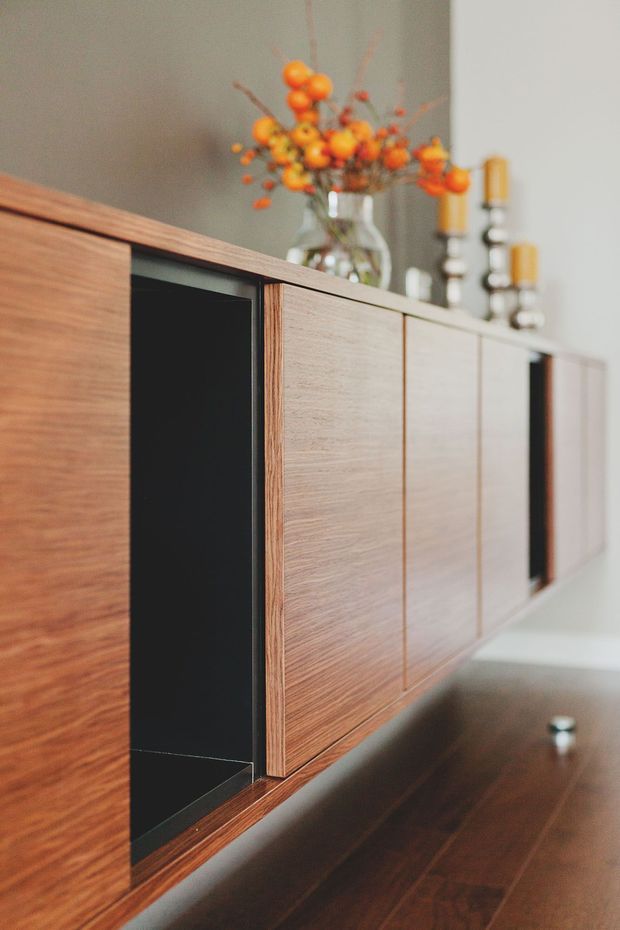 Close-up interior detail of a mid-century-inspired timber sideboard or built-in cabinet, featuring warm wood veneer fronts, contrasting black inset panels, and a dark timber top. Decorative glass vase with orange foliage and candleholders sit on the cabinet against a muted grey wall.