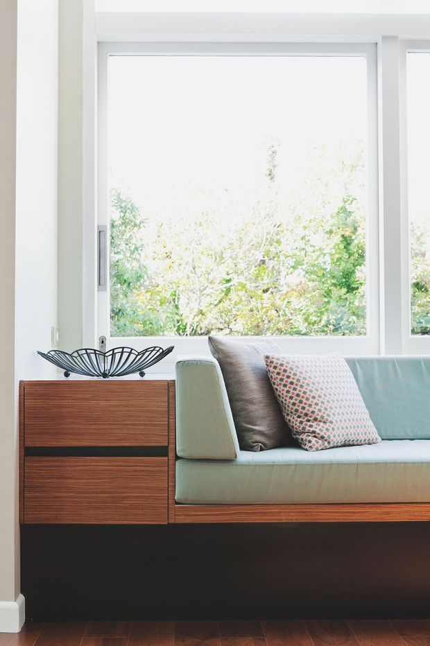 Bright interior detail of a built-in living room window seat with pale blue-green upholstered seating, patterned cushions, and integrated dark timber cabinetry. A large white-framed window overlooks leafy greenery, while warm timber flooring and a sculptural black wire bowl add contrast. The design has a clean mid-century-inspired character.