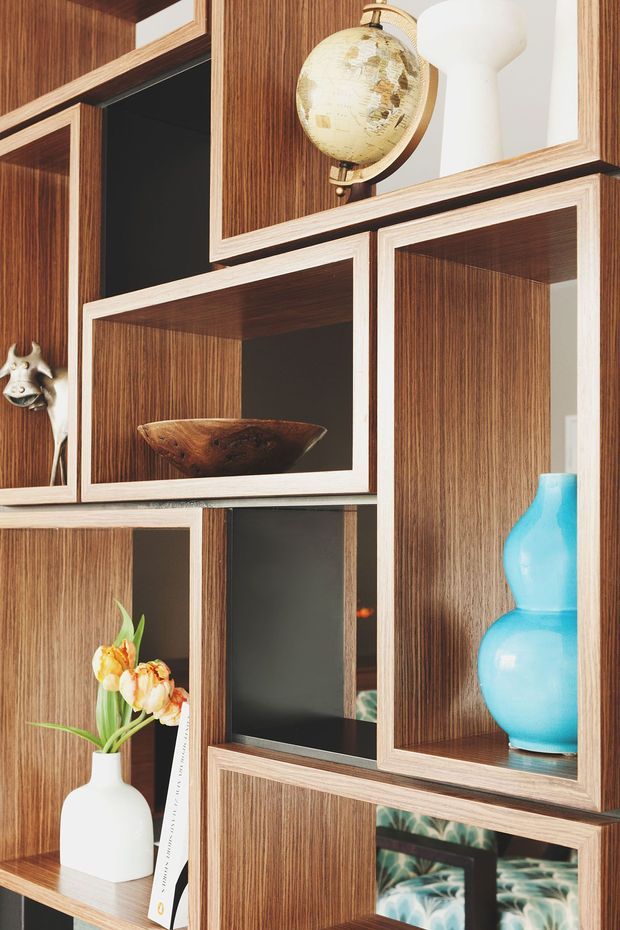 Close-up interior detail of a sculptural built-in display shelving system composed of staggered rectangular compartments. The joinery has richly grained dark timber veneer with pale contrasting trim and dark recessed interiors. Shelves display a globe, white and turquoise ceramic vases, a wooden bowl, flowers, and decorative objects, creating a curated mid-century-inspired feature wall.