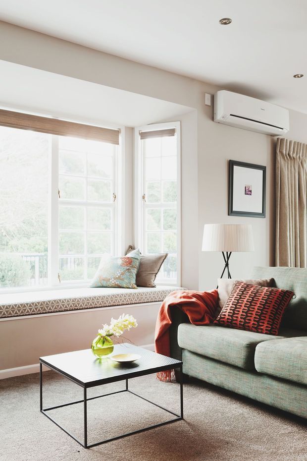 Bright, refined living room with a large window seat, tall divided-light windows, neutral walls, beige carpet, and soft taupe curtains. A pale green upholstered armchair, black rectangular coffee table, floor lamp, patterned cushions, and a red throw create understated colour accents. A wall-mounted split-system air conditioner is visible above a framed artwork.