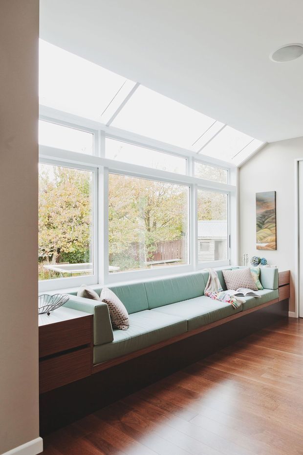 Bright, refined living room with a long built-in window seat and integrated timber cabinetry beneath a large bank of white-framed windows and clerestory glazing. The room has warm timber flooring, pale walls, a muted green upholstered bench, patterned cushions, and a small artwork. Abundant natural light creates an airy, calm atmosphere.