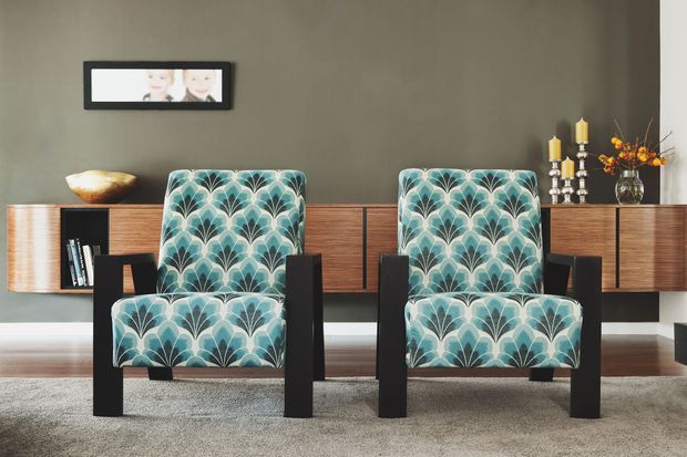 A refined living room vignette featuring two matching upholstered armchairs in a teal, cream, and grey scalloped pattern, positioned in front of a long dark timber wall-mounted sideboard. The room has a muted taupe wall, light carpet, minimalist framed artwork, decorative objects, and warm metallic candleholders.