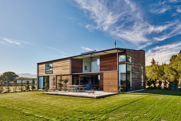 Exterior view of a contemporary two-storey home with a rectilinear flat-roof form, extensive glazing, dark timber horizontal cladding, and a covered outdoor terrace opening onto a broad lawn. The house is set in a landscaped suburban or semi-rural setting with distant hills visible.