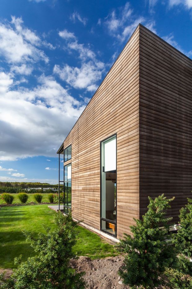 Exterior architectural photograph of a tall, sharply angled modern building clad in horizontal timber boards. Narrow vertical glazing and a glazed corner articulate the facade, while landscaped garden planting and lawn surround the structure. The image is taken from a low angle beneath a bright blue sky with scattered clouds.