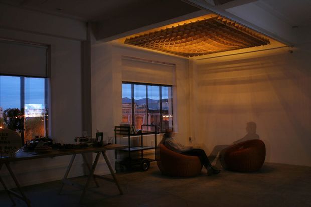 Dimly lit contemporary interior lounge or office space with large windows overlooking an urban landscape. Two rounded lounge chairs sit beneath a dramatic suspended timber lattice ceiling feature with warm integrated lighting. A long worktable and shelving occupy the left side, while exposed concrete-like surfaces and roller blinds contribute to an industrial-contemporary atmosphere.
