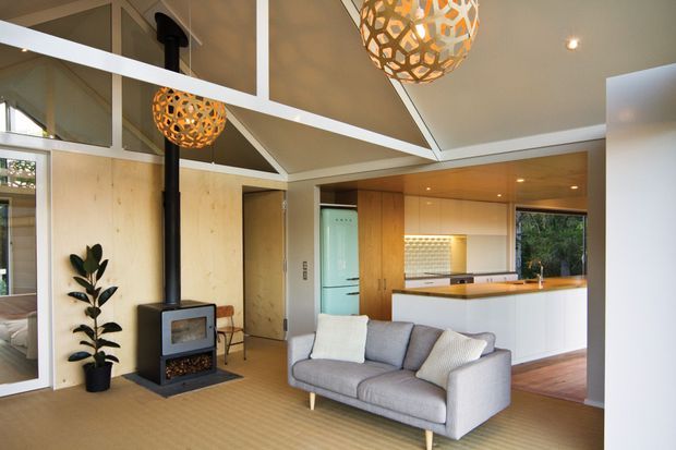 Open-plan living and kitchen interior with a warm timber-lined fit-out, a central black wood-burning stove, grey sofa, polished timber flooring, and a compact kitchen with timber cabinetry, white lower cabinets, and a timber benchtop. The room has a distinctive vaulted geometric ceiling with exposed white structural beams and decorative pendant lights.