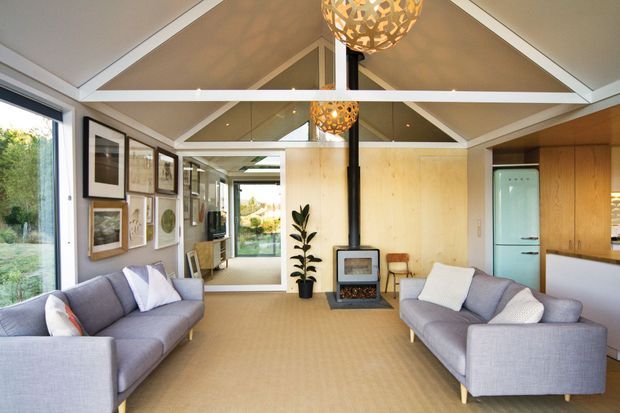 Bright open-plan living room with two grey sofas facing each other, a central freestanding wood-burning stove, pale timber wall panelling, large glazed openings, and a vaulted ceiling with exposed white framing. Decorative pendant lights, framed artwork, and a potted plant add character. The room opens toward a sunlit exterior view and has a calm, contemporary coastal feel.