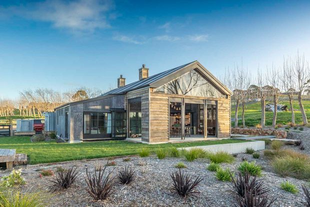 Single-storey contemporary rural-style house with a prominent pitched gable roof, dark metal roofing, weathered timber cladding, extensive glazing, and a landscaped gravel garden. The building appears to sit in an open rural or semi-rural setting, with lawn, planted borders, and leafless trees surrounding it.