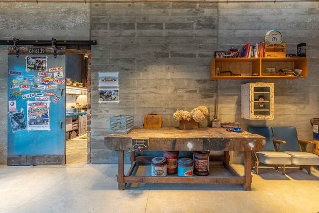 An industrial-style interior with a grey concrete-block wall, polished concrete floor, and a large rustic timber worktable or console. A vintage blue sliding door covered in stickers opens to an adjoining space, while timber wall shelving, framed artwork, books, paint tins, and mid-century-style chairs add character. The room has warm ambient lighting and a workshop-inspired, eclectic atmosphere.